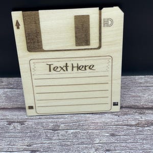 May include: A wooden floppy disk-shaped item with the words "Text Here" engraved on the front. The design includes a dark brown top section with an arrow and a rectangular cutout. The item is on a wooden surface.