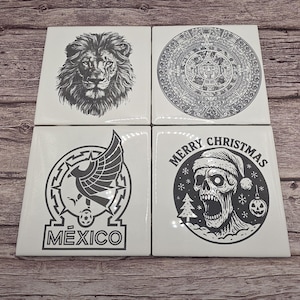 May include: Four white square ceramic coasters with black designs. The designs include a lion's face, an Aztec calendar, a soccer emblem with the word "MEXICO", and a skull wearing a Santa hat with the words "MERRY CHRISTMAS".