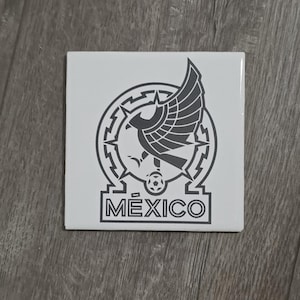 May include: Square white tile featuring the emblem of Mexico. The emblem includes a stylized eagle with outstretched wings, a soccer ball, and the word "MÉXICO" in a bold font. The design is in shades of gray.