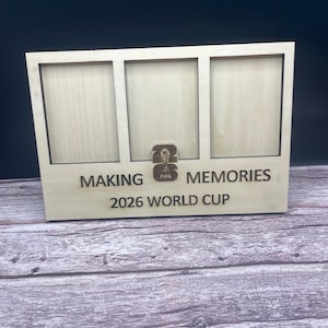 May include: A light-colored wooden triple photo frame featuring three rectangular openings. The frame is engraved with the text "MAKING MEMORIES" and "2026 WORLD CUP," and includes a small central emblem with the number 26 and the FIFA logo.