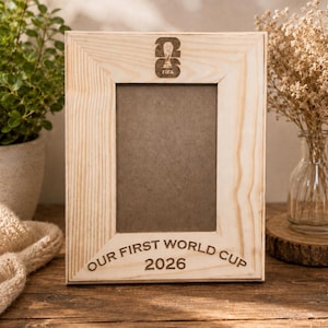 May include: A rectangular light-colored wooden picture frame with a visible grain. The top center features an engraved FIFA World Cup trophy logo. The bottom center is engraved with the text "OUR FIRST WORLD CUP 2026" in dark lettering.