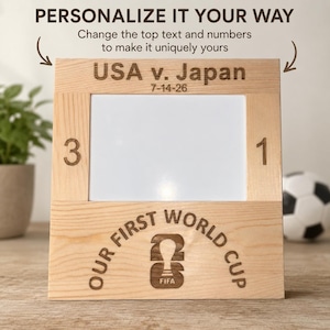 May include: A light-colored wooden picture frame with engraved text reading USA v. Japan 7-14-26, the numbers 3 and 1 on the sides, and the text Our First World Cup with a FIFA logo at the bottom. The frame features a central white rectangular insert.
