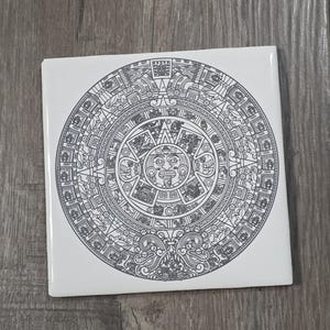 May include: Square white tile featuring a detailed black and gray illustration of the Aztec calendar. The circular design includes intricate patterns and a central sun face. The tile is likely ceramic or a similar material.