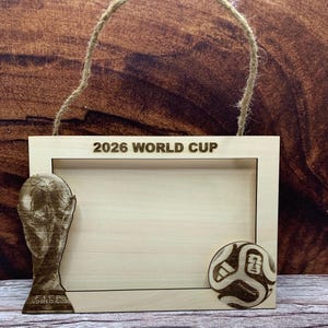 May include: A rectangular light-colored wooden picture frame with a jute twine hanger. The top edge features the text 2026 WORLD CUP. The left side has a laser-engraved FIFA World Cup trophy, and the bottom right corner has a soccer ball graphic.