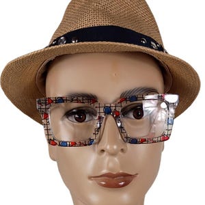 May include: A straw fedora hat with a black band and silver studs, paired with square-framed eyeglasses. The eyeglasses have a clear lens and a patterned design with red, blue, and black shapes.