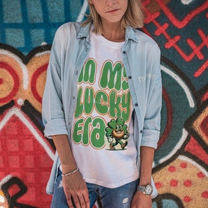 May include: A white t-shirt with a green shamrock graphic and the text "In my lucky era". The shirt is being worn by a person standing in front of a colorful graffiti wall.