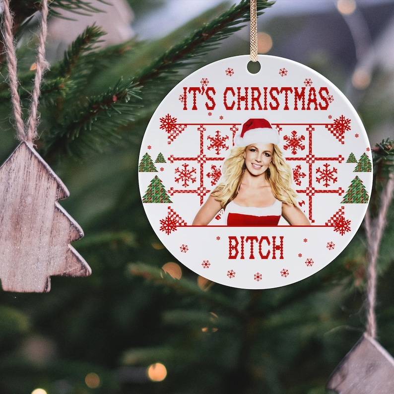 May include: A white ceramic Christmas ornament with a red and white pixelated design. The ornament features a photo of a woman wearing a Santa hat and a red dress. The text "It's Christmas Bitch" is printed on the ornament.