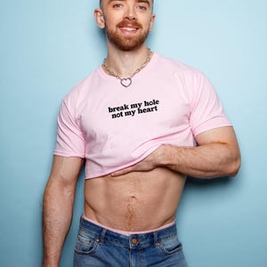 May include: A light pink crop top with the text "break my hole not my heart" in black. The top is paired with blue denim jeans. A silver chain necklace with a heart pendant is also visible.