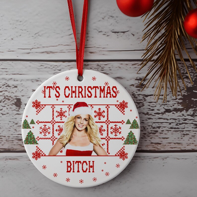 May include: White ceramic Christmas ornament with a red and white pixelated design. The ornament features a photo of a woman wearing a Santa hat and a red and white dress. The text "It's Christmas Bitch" is printed on the ornament.