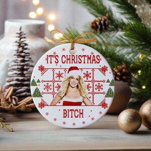 May include: A white ceramic Christmas ornament with a red and white pixelated design. The ornament features a photo of a woman wearing a Santa hat and a red dress. The text "It's Christmas Bitch" is printed on the ornament.