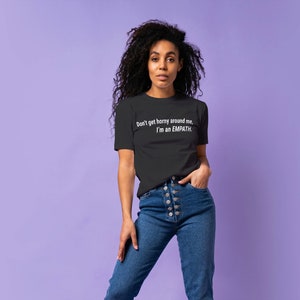 May include: A woman wearing a black t-shirt with the text "Don't get horney around me, I'm an EMPATH."  She is wearing blue jeans and has curly dark hair.