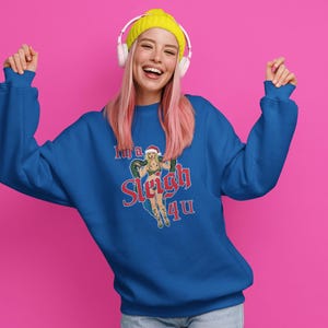 May include: A royal blue sweatshirt with the text "I'm a Sleigh 4 U" and a graphic of a woman in a Santa hat. The person in the image is wearing a yellow beanie and headphones, with pink hair, and is smiling.
