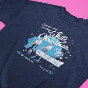 May include: A navy blue sweatshirt with a graphic from the film "White Christmas." The design includes the film's title, the year 1954, and an illustration of two men. The text also mentions "The Haynes Sisters" and "The Columbia Inn."
