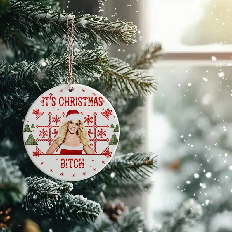 May include: White ceramic Christmas ornament with a red and white knitted pattern. The ornament features a photo of a woman wearing a Santa hat and a red dress. The text "It's Christmas Bitch" is printed on the ornament.