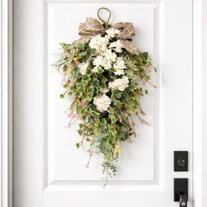 Op de afbeelding: Een waterval van bloemen met witte bloemen, groene bladeren en roze accenten, bekroond met een bruine strik met patroon. De bloemstuk hangt aan een groene lus, geschikt voor deur- of wanddecoratie.