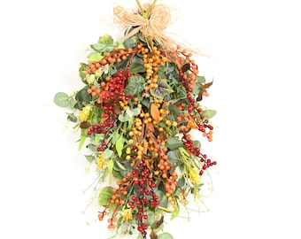 May include: A decorative teardrop-shaped wall hanging featuring a mix of green foliage, yellow and red berries, and dried leaves. The arrangement is tied at the top with a natural-colored raffia bow and includes a small green loop for hanging.