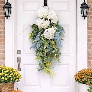Op de afbeelding: Een witte voordeur versierd met een trapsgewijze bloemstuk van witte hortensia's, blauwe accenten en groen. De deur wordt geflankeerd door bakstenen muren en zwarte lantaarns. Pompoenen en potchrysanten maken de herfstdecoratie compleet.