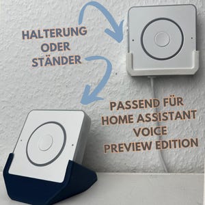 Op de afbeelding: Een wit vierkant apparaat met een centrale cirkelvormige knop, gemonteerd in een witte houder, en een apart apparaat in een donkerblauwe standaard. Tekst op de muur: "HALTERUNG ODER STÄNDER" en "PASSEND FÜR HOME ASSISTANT VOICE PREVIEW EDITION."