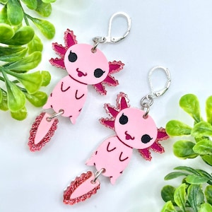 May include: Pink axolotl earrings with silver hooks. The axolotls have pink bodies, black eyes, and red glitter accents.