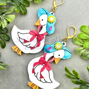 May include: A pair of white goose earrings with blue hats, pink bows, and yellow flowers. The earrings are made of resin and have gold hooks.