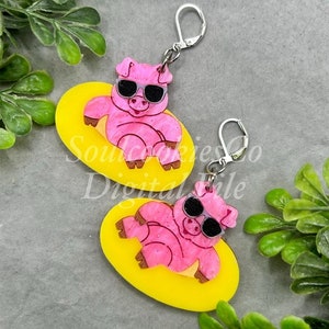 Puede incluir: Un par de pendientes de cerdo rosa con gafas de sol, cada cerdo está tumbado en un flotador amarillo ovalado. Los pendientes tienen ganchos plateados.
