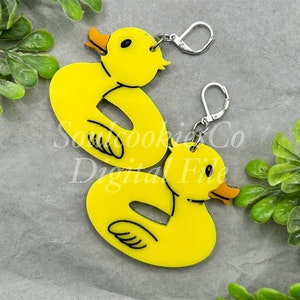 Puede incluir: Un par de pendientes de pato de goma amarillo con ganchos plateados. Los patos están en posición de natación con la cabeza girada hacia un lado.