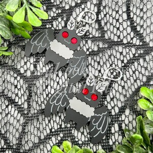 May include: A pair of black bat earrings with red eyes and white fur. The earrings are made of acrylic and have silver hooks.
