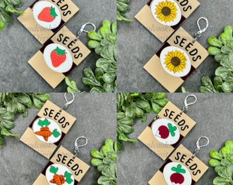 Könnte beinhalten: Ein Paar Hängeohrringe mit einem braunen rechteckigen Anhänger, auf dem das Wort "SEEDS" in Schwarz gedruckt ist. Jeder Anhänger zeigt eine farbenfrohe Illustration einer Erdbeere, einer Sonnenblume, einer Karotte oder einer Rübe.