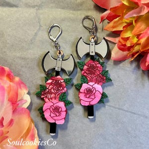 Può includere: Un paio di orecchini unici con un design a forma di ascia argentata e un manico nero, adornati con rose rosa e foglie verdi. Gli orecchini hanno chiusure a leva argentate. Il testo "SoulcookiesCo" è visibile.