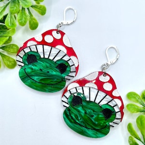 Puede incluir: Un par de pendientes con forma de seta roja y blanca con una cara de rana verde. La seta tiene lunares blancos y una franja blanca alrededor de la parte superior.