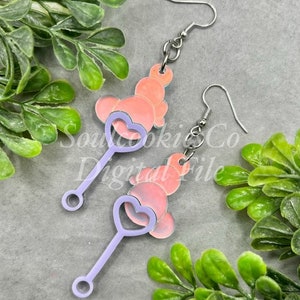 Puede incluir: Un par de pendientes colgantes con un dije en forma de corazón rosa y blanco con un palo morado adjunto. Los pendientes son de metal y tienen un gancho plateado.