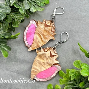 Può includere: Un paio di orecchini a conchiglia rosa e oro con ganci d'argento. Le conchiglie sono dettagliate con un motivo marmoreo e hanno un interno rosa.