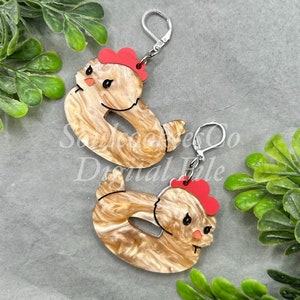 May include: A pair of earrings featuring a brown and white marbled chick design with red accents. The earrings are made of acrylic and have silver hooks.