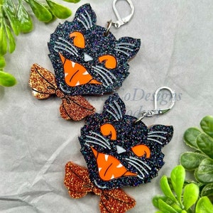 May include: A pair of black glitter cat face earrings with orange glitter bow ties. The cat faces have white eyes and a wide, toothy grin.