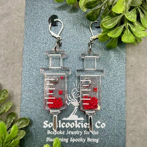 May include: A pair of silver earrings shaped like syringes filled with red glitter. The earrings are on a silver background with the text "Sollcookie Co. Bespoke Jewelry for the Discerning Spooky Being".