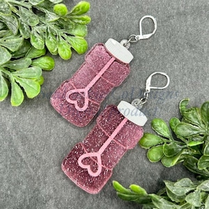 Puede incluir: Pendientes de botella de burbujas de purpurina rosa con ganchos plateados. Las botellas tienen una tapa blanca y un diseño de corazón rosa.