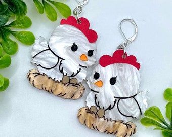 May include: A pair of white and red chicken earrings with a nest design. The chickens have black eyes and orange beaks.