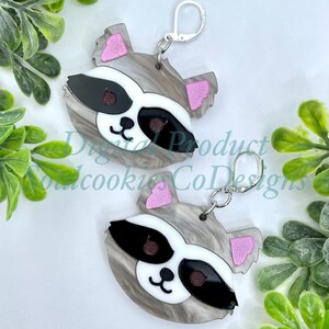 May include: A pair of raccoon face earrings with pink ears, black eyes, and a white nose. The earrings are made of gray and white marbled acrylic and have silver hooks.
