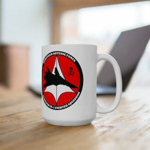 May include: White ceramic mug with a black and red design. The design features a stylized fighter jet, a skull and crossbones, and the text "Robotech Defense Force VF-1 Fighter Squadron".