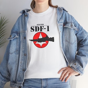 Puede incluir: Camiseta blanca con un círculo rojo y un gráfico de nave espacial negra. El texto "Deck Crew SDF-1" está impreso en negro sobre el gráfico.
