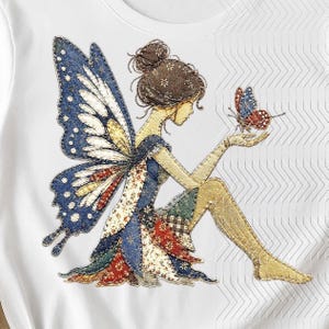 Puede incluir: Una camiseta blanca con un gráfico bordado detallado de un hada con grandes alas estampadas en azul y blanco. El hada tiene el cabello oscuro recogido en un moño y está sentada mientras sostiene una pequeña mariposa roja y azul en una mano. El diseño utiliza varias texturas y patrones de tela en tonos azul, rojo, blanco y marrón para crear una apariencia de capas estilo patchwork.