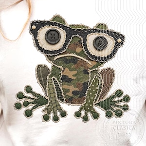May include: A white t-shirt featuring a fabric applique of a frog with a camouflage pattern body. The frog wears black glasses made from fabric and black buttons. The limbs are crafted from green fabric with polka dot and plaid patterns.