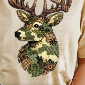 May include: A light beige t-shirt featuring a graphic of a deer head. The deer is rendered in a patchwork style with various green, brown, and tan camouflage and plaid fabric patterns. It has large brown antlers and a small wooden button detail.