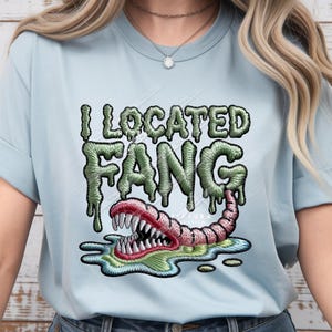 May include: A light blue t-shirt featuring a graphic design with the text I LOCATED FANG in a green, dripping, embroidered-style font. Below the text is a pink, worm-like creature with sharp white teeth emerging from a green puddle.