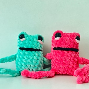May include: Two crocheted frog plushies, one teal green and one hot pink, with black eyes and mouths. The frogs are sitting side by side on a white surface.