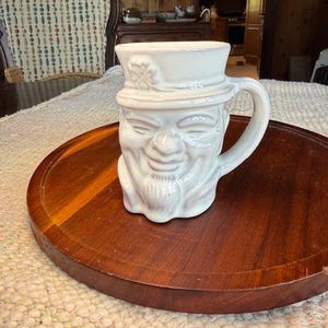 May include: A white ceramic mug with a face design, featuring a hat and a smiling face with a beard. The mug is on a round, wooden tray.