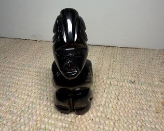 May include: A small, black carved statue of a figure with a stylized face and body. The statue has a glossy finish and appears to be made of a solid, dark material. The figure is seated and has a detailed head with a prominent forehead.