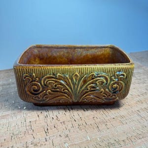 Puede incluir: Un plato rectangular de cerámica con un esmalte marrón y dorado. La parte delantera del plato presenta un diseño floral en relieve. El plato tiene una superficie texturizada y probablemente se utiliza con fines decorativos.