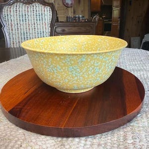 May include: A large, yellow mixing bowl with a speckled blue and white pattern. The bowl sits on a round, dark brown wooden serving tray. The bowl is centered on a white, textured surface.