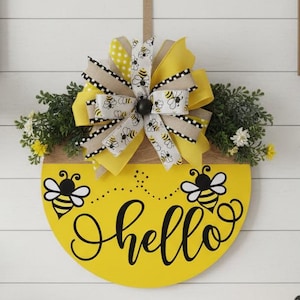 May include: A yellow wooden sign with the word "hello" in cursive, decorated with bee graphics and a large bow. The bow features yellow, white, and tan ribbons. The sign is adorned with greenery and small yellow flowers, creating a welcoming aesthetic.